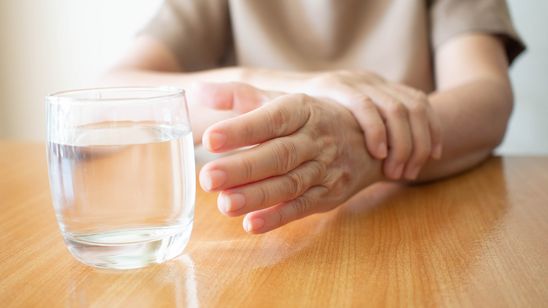 Frau mit Parkinson versucht, ein Wasserglas zu greifen und muss dabei mit der anderen Hand ihre Greifhand festhalten